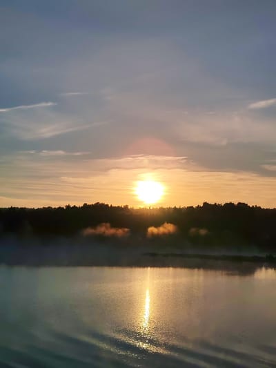 This image captures a sunrise or sunset over a body of water in Kabanovo, Russia. The sun, a prominent bright yellow-orange sphere with a noticeable lens flare, is positioned low on the horizon, slightly to the left of the image's vertical center. The sky exhibits a gradient, transitioning from a light blue-grey at the top to warm orange and yellow tones closer to the horizon, with several thin, elongated clouds scattered across it. A dark, dense silhouette of a forest or tree line forms the horizon, dividing the sky from the lower elements of the scene. Below this tree line lies a calm body of water, likely a lake or wide river, which strongly reflects the sun's light, creating a bright, shimmering vertical path on its surface. Wisps of atmospheric mist or fog, illuminated by the sun's glow with hints of orange, are visible just above the water's surface, particularly in front of the dark tree line. No individuals, man-made structures, or vehicles are discernible within the frame.