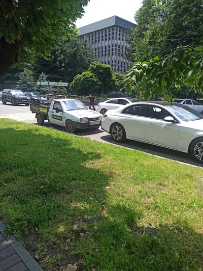 In a scene set in Atabe, Georgia, a row of parked cars lines a grassy roadside next to a sidewalk. The time of day appears to be late morning or early afternoon, indicated by the bright sunlight and distinct shadows cast on the grass. The weather is fair and sunny, with lush green trees flanking the area and a large, modern building visible in the background.
Among the vehicles, a white sedan with a sleek design is prominent in the foreground, parked parallel to the road. Behind it, a white utility truck with a flatbed is parked, displaying visible text on its side. The text appears to be in Cyrillic script and reads "ОКНА БЕРТА" (OKNA BERTA), suggesting a business related to windows. Further down the line, other cars are visible, including a dark-colored SUV. A person dressed in black is seen standing near the utility truck, and another individual in an orange jacket is visible further down the street. The overall setting suggests a typical urban or suburban environment in Georgia during a sunny day.