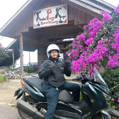A man wearing a helmet and gloves sits astride a dark green Yamaha scooter on a paved surface. He is smoking a cigarette and waving with his left hand. Behind him is a wooden building with a sign reading "Kedai Langit Kopi BurniteLong" in black, red, and white lettering. To the right of the building is a large bush of bright pink bougainvillea flowers. The scene is located in the city of Panjimulia Satu, Indonesia. The backdrop includes a slightly elevated view and an additional structure with a thatched roof.