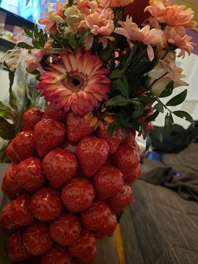 The scene depicts an indoor environment in Oberhausen, Germany, featuring a floral and fruit arrangement. The primary subject is a vertical bouquet composed of a dense cluster of approximately 30-40 bright red, glossy strawberries forming the base, topped by a mixed assortment of fresh flowers. The flowers include several peach and pink chrysanthemums, a prominent Gerbera daisy-type flower with bi-colored red-pink and cream petals and a dark central disc, and several pale pink/white rose-like blossoms, all interspersed with green foliage. The arrangement is illuminated from the front. In the blurred background to the left, a television screen displaying a bright blue and white image is partially visible above indistinct furniture. To the right, a textured grey-brown fabric surface, likely a sofa or bed, is visible, with a crumpled dark fabric object resting upon it. No individuals are present in the image.