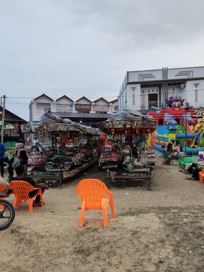 This image depicts an outdoor amusement area on a cloudy day in Peurupok, Indonesia. In the foreground, two bright orange plastic chairs sit on a dirt and gravel surface. To the left, a motorcycle wheel is visible. In the mid-ground, a colorful carousel with small ride-on vehicles, decorated with lights and imagery, is operational. Several people, including adults and children, are interacting with the carousel. To the right of the carousel, a large inflatable bounce house, shaped like a robot and featuring a cartoon bus, is set up. More people are seated on green plastic chairs near the bounce house. Behind the amusement area, several commercial buildings with modern architecture line the street. Signs on these buildings indicate businesses such as a dental practice ("PRAKTEK GIGI"), a studio ("Etc Studio"), and a hair salon ("LONDON & HAIRCUT"). The overall scene captures a community gathering or local event with entertainment options.