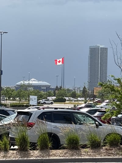 The image shows a Canadian flag flying in the wind in a parking lot in Vaughan, Canada. In the background, there are several tall buildings and a large domed structure, likely a shopping mall or entertainment venue. The parking lot is filled with cars, and there are trees and grass in the foreground. The sky is overcast, suggesting it might rain.