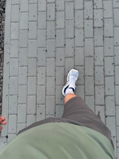 A first-person perspective shot shows a person walking on a paved surface in Horenychi, Ukraine. The ground is composed of rectangular gray pavers arranged in a running bond pattern. To the left of the pavers, there is a strip of dark gravel. The person is wearing a white athletic shoe with black accents, white socks, dark gray shorts, and a light green shirt or jacket. The right leg and foot are visible, mid-stride, with the left leg out of frame. A hand with visible fingers is also partially visible on the left side of the frame. The lighting suggests an outdoor, daytime setting.