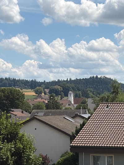 The image shows a daytime view of Lotzwil, Switzerland, focusing on a residential area with a backdrop of a tree-covered hill and a blue sky filled with white, puffy clouds. In the foreground, several houses with tiled roofs are visible, some partially obscured by lush green trees. The houses exhibit typical European architectural styles, with visible chimneys and windows.
Further into the scene, a distinct white church tower with a clock face and a steeple stands out amongst the surrounding buildings. The church is a prominent landmark, suggesting it is likely the village church of Lotzwil. Beyond the residential area and the church, the landscape transitions to rolling hills, with fields of golden crops interspersed with dense green forests. The overall impression is of a peaceful, rural village scene. No people are visible in the image.