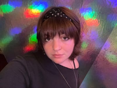 A young woman with brown hair and bangs, wearing a black and white polka dot headband, is positioned upside down in the frame. She has a nose ring and is looking directly at the viewer. She is wearing a black top with thin straps. The background has an iridescent sheen, reflecting rainbow colors and creating a holographic effect. Part of the background is a large, grey triangle.