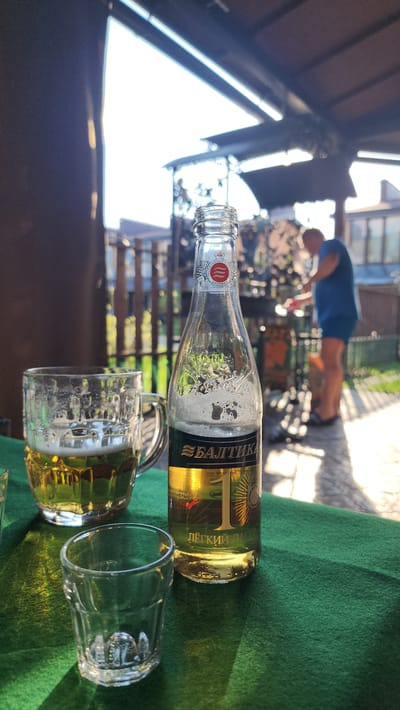 The image depicts a scene in what appears to be an outdoor setting. In the foreground, there's a bottle of Baltika beer (possibly Baltika 1), a partially full beer mug, and an empty glass. These are sitting on a green surface, possibly a tablecloth or outdoor table covering.

In the background, a person wearing a blue shirt and gray shorts is seen performing what seems to be push-ups. The background environment also includes elements of an outdoor structure or gazebo-like area with wooden supports and a partially obscured landscape beyond.

Given the writing on the bottle, specifically mentioning "Baltika," and the Cyrillic text, it seems likely the location is in Russia or a country where Russian is spoken. Given the prompt information, the location is in the city of Рыбхоз, Russia.