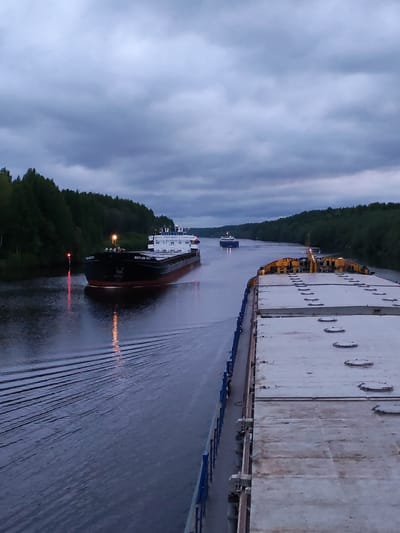 The image depicts a waterway with two ships passing each other in Александровское, Russia. The sky is overcast. On the left, a black and red cargo ship, "Volgo-Don 5073," is underway, creating visible wakes. A red marker buoy is visible near the edge of the waterway. On the right, a flat-topped barge is docked, showing its deck with metal features. A second ship, further in the distance, is also navigating the waterway. Dense forest lines both sides of the canal.