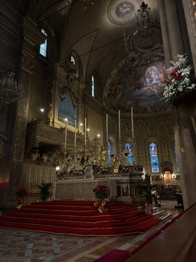 This image depicts the interior of the Basilica Cattedrale di San Giorgio Martire (Ferrara Cathedral) in Ferrara, Italy, specifically facing the main altar and apse from the nave. The scene is illuminated by dim ambient light, supplemented by artificial spotlights and lit candles.
In the foreground, five red carpeted steps with gold trim descend from a raised platform. Two potted plants with red flowers, wrapped in gold foil, flank the steps. The visible floor is patterned marble.
At the center, the main altar stands on a platform, featuring a white, carved altar frontal. It is adorned with ornate gold and silver candelabras holding tall white candles, some actively lit. Behind the altar, the apse wall is richly decorated with gold relief work and multiple stained-glass windows displaying figures in blue, red, and yellow tones. Above the apse, a large, detailed fresco, likely depicting the Last Judgment, covers a significant portion of the curved wall, featuring numerous figures in a dynamic composition. To the right of this fresco, a large arrangement of white lilies and red flowers rests on a stand.
On the left side of the image, an elaborate pipe organ, predominantly gold and white with visible silver pipes, is mounted on a decorated balcony structure. The surrounding walls feature intricate golden architectural details.
The vaulted ceiling above includes a circular fresco depicting the Agnus Dei (Lamb of God) holding a banner. A large, multi-tiered crystal chandelier hangs from the ceiling towards the right side of the frame. No individuals are present in the scene.