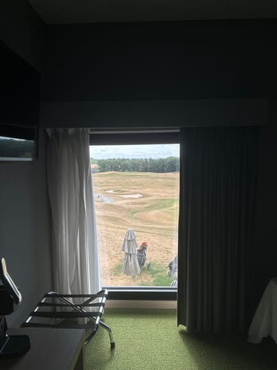 The image shows a room with a large window overlooking a golf course. The room has a luggage rack, a desk with a black object on it, and green carpet. The window is partially covered by white curtains on the left and dark curtains on the right. Outside the window, a golf course is visible with rolling hills, sand traps, and trees in the distance. There is a golf bag with an umbrella and a golf cart in front of the window. The scene appears to be during the day, with overcast skies. The location is the city of Méan, Belgium.