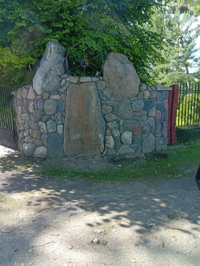 A rustic stone wall, built from a variety of irregularly shaped stones in shades of gray, brown, and blue, stands as a central feature in the scene. Integrated into the wall are several large, rough-hewn rocks, one of which on the left is significantly larger and more imposing than the others. At the peak of the wall, between two of these larger rocks, a metal spoked wheel is prominently displayed. A tall, rectangular stone slab is set vertically into the face of the wall, centrally located, providing a stark contrast to the surrounding stonework.
To the left of the stone wall, a black metal fence extends outwards, partially visible. To the right, a tall red wooden post stands next to a similar black metal fence, suggesting a gateway or entrance. Behind the wall, lush green trees and pine trees create a natural backdrop, with sunlight filtering through the leaves, casting dappled shadows on the ground. The foreground consists of a dirt and gravel path, also marked with shadows from the overhead foliage.
The scene appears to be set outdoors during the daytime, likely on a sunny day given the distinct shadows. The overall impression is one of a natural, somewhat rural setting, possibly a park, garden, or the entrance to a property. There are no people visible in the image, and no text is present. The atmosphere feels calm and tranquil, with the stone wall and natural elements dominating the visual narrative. The location is Micaičiai, Lithuania.