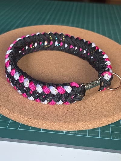A braided bracelet with black, pink, and white cords, accented with metallic gold and blue threads, is laid flat on a round cork coaster. The bracelet features a silver clasp and a silver D-ring. The coaster rests on a green cutting mat with white grid lines. The background is blurred, suggesting an indoor setting. There are no people present. The location in the city of Coslada, Spain, cannot be determined from the image.