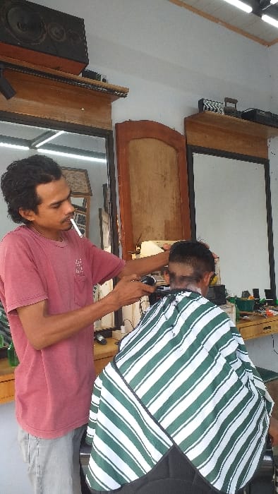 The image displays a barbershop scene in Serdang, Indonesia, where a male barber is actively cutting a customer's hair. The barber, with dark wavy hair, is attired in a red short-sleeved t-shirt and light-colored trousers, holding an electric hair clipper in his right hand. A lit cigarette is present in his mouth. The customer, viewed from behind, is seated and covered by a green and white striped barber cape, revealing a partially faded haircut. The background consists of white walls, two wooden-framed mirrors, and wooden shelves displaying various barbering tools and supplies. A large black speaker is visible on the top shelf to the left, and exposed fluorescent light fixtures are mounted on the ceiling.