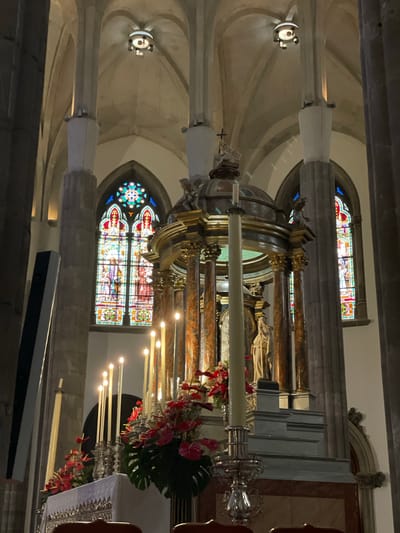 The image captures the elaborate interior of a church or cathedral, viewed from a side angle towards a richly decorated central structure. In the foreground and midground, an ornate processional float or altar, crafted from golden and dark wood with classical columns, is prominently displayed. It features statues of religious figures, several tall white candles in ornate candelabras (some lit), and vibrant floral arrangements of red flowers and large green Monstera leaves. A thick, unlit white candle lies horizontally across the midsection of the float.
In the background, two large, arched stained-glass windows, filled with colorful depictions of religious scenes, flank the central structure. Tall, light-colored fluted columns support a vaulted ceiling, typical of historical ecclesiastical architecture. Spotlights are visible on the ceiling and columns, providing illumination. On the right, a white cloth with intricate silver embroidery is partially visible.
The lighting within the church is dim, creating a solemn atmosphere, with the candles and stained glass serving as primary light sources. No people are present, but the arrangement strongly suggests a religious ceremony, a special event, or a processional display within the sacred space. A faint label with the numbers "177_000_177_379" is visible on a white strip attached to one of the columns. The setting is consistent with a church in San Cristóbal De La Laguna, Spain.