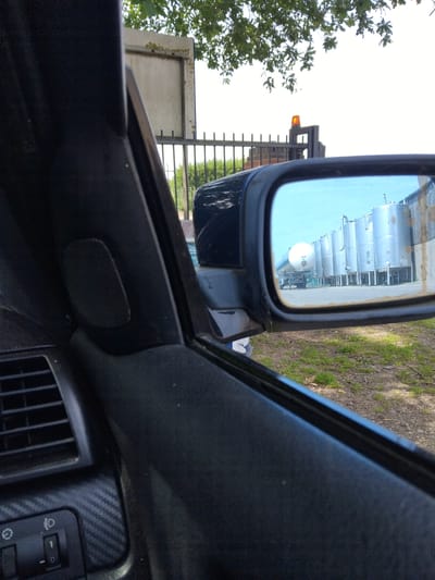 The image is taken from inside a vehicle, looking towards the side mirror. The side mirror reflects a row of tall, cylindrical silver storage tanks, likely part of an industrial facility. In the foreground, partially visible, is the interior of the car, including part of the dashboard, a speaker, and a vent. Outside the car, a metal fence and some trees are visible. The sky is bright and overcast. There are no people clearly visible in the image. The location in the city of Stafford, United Kingdom, is not specified or discernible from the image itself.