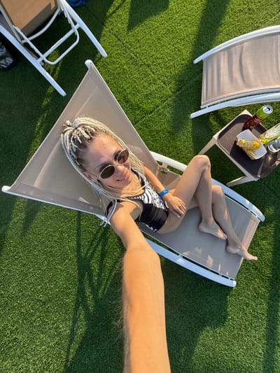 The image shows a woman with blonde braids relaxing on a lounge chair on a grassy area, possibly a park or resort, under a bright sun. She is wearing a black and white patterned swimsuit and sunglasses. A small table next to her holds a drink and some food. The perspective suggests the photo was taken from a high angle, possibly by someone using a selfie stick. The environment appears to be outdoors with artificial turf and shadows from unseen objects.