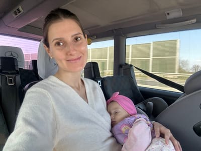 The image captures an interior view of a vehicle, depicting an adult female holding an infant, purportedly while traveling on a highway in Grodziec, Poland.
The adult female, with light skin and dark hair pulled back, wears a light-colored, possibly off-white, knitted v-neck sweater. She is positioned towards the left side of the frame, looking directly at the viewer with a slight smile. She holds an infant cradled against her left side. The infant, wearing a pink headwrap, a purple long-sleeved top with a rainbow motif, and a light pink lower garment, is asleep with eyes closed and head resting against the adult's arm. The infant is partially secured within a grey child car seat, visible to the right of the adult.
The vehicle interior features dark grey upholstered seats and headrests, at least two of which display a Mercedes-Benz logo. A black seatbelt is visible across the mid-ground. The overhead panel shows light-colored ceiling material with a handle and a light fixture. Through the vehicle's windows, a multi-lane highway is visible, bordered by tall, light-colored sound barrier walls featuring a horizontal grid pattern. Beyond the barriers, bare trees are visible under a clear, blue sky, indicating an outdoor environment, likely a spring or late winter day. The scene suggests a journey in progress.