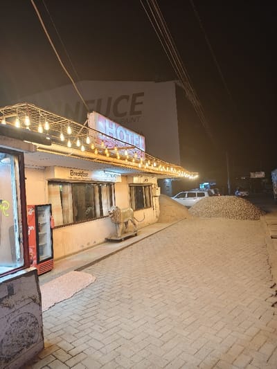 A street view at night features a small hotel or restaurant with "Hotel" prominently displayed on its sign. String lights illuminate the facade and a lion statue stands near the entrance. Next to the building are piles of sand or gravel. Cars and faint signs of other structures are visible in the background under a dark sky with electrical wires crossing overhead.