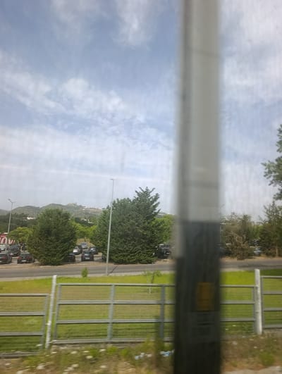 The image depicts an outdoor scene from a moving vehicle, likely a train, passing through Palmela, Portugal. A blurred vertical pole occupies the foreground, obscuring a portion of the view. Beyond the pole, a road with parked cars is visible, bordered by a green grassy area with a metal fence. Mature trees, some evergreen and others with bare branches, are scattered throughout the landscape. In the background, rolling hills are covered with vegetation, and a distant castle or fortress stands atop one of the highest points. The sky is partly cloudy with a light blue hue.