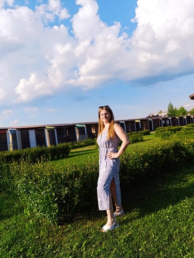 A full-body shot captures a woman standing on a grassy area with lush green bushes in the foreground and a row of small, wooden structures in the background. The sky above is a vibrant blue, dotted with fluffy white clouds. The woman, with long, light brown hair, is wearing a light blue dress with a white floral pattern and a thigh-high slit. She has on sunglasses pushed up on her head, a ring on her left hand, and is posing with her left hand on her hip. Her white sandals are visible on her feet. The structures in the background appear to be beach or changing huts, each with a dark wooden facade and a colorful trim along the roofline, suggesting a seaside or recreational location, possibly in Ivanovskoe, Russia. The lighting indicates it is daytime, likely late afternoon, with the sun casting shadows on the grass.