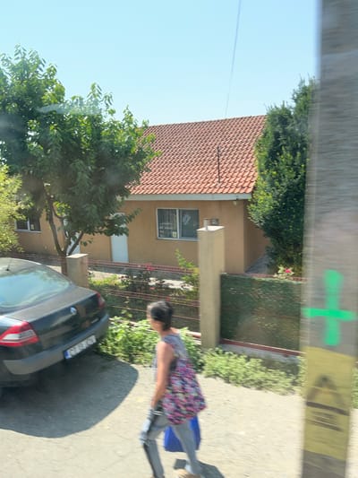 This image captures a residential scene in Movilița, Romania, on a clear, sunny day. In the foreground, a woman is walking away from the camera, wearing a colorful floral top and gray pants, carrying a blue bag. To her left, a dark-colored car is parked, its rear end visible. Behind the car and the woman is a low fence with red posts and green mesh, separating the property from the street. Beyond the fence, a house with a light orange facade and a red tile roof is visible. Two windows with white frames are prominent on the facade, and a white garage door is also present. Lush green trees and bushes surround the house and fence, with some red flowers blooming near the fence. A light pole with green markings is visible on the right side of the frame, indicating urban infrastructure. The sky is a bright, clear blue.