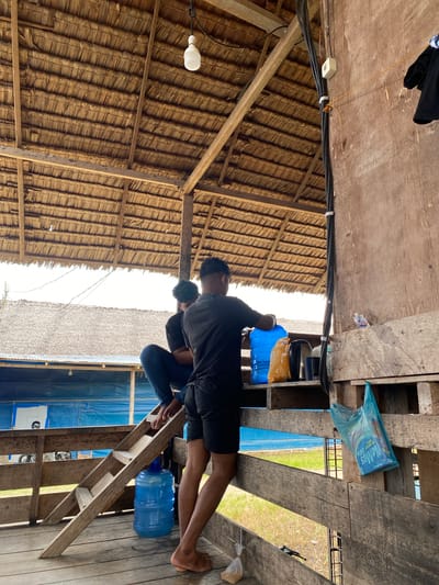 The image captures an outdoor scene under a thatched roof structure. Two individuals are present. One person, wearing a black t-shirt and shorts, stands by a table, reaching towards a large blue water cooler and an orange plastic bag. Another person, in a black t-shirt and jeans, is seated to the left, partially obscured. The environment appears rustic, with visible wooden beams and planking. A wooden ladder leans against a railing. Various objects, including a kettle, a plastic bag with items, and a small plastic packet on the floor, are scattered around. The background reveals a blue tarp-covered structure and distant foliage. The location is identified as Monjeureujak, Indonesia.