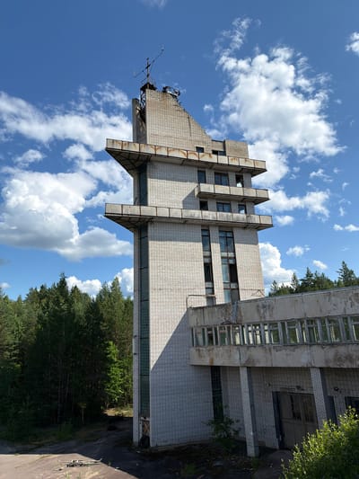 This image shows an abandoned, multi-story building in Nikolsky, Russia, set against a backdrop of a blue sky with white clouds and a dense forest. The building is constructed of light-colored bricks and features several balconies with metal railings. The architecture is angular, with some sections appearing to jut out. The windows are numerous, and many of them appear broken or boarded up. At the top of the tallest section of the building, there is a structure resembling an antenna or communication tower. The ground around the building is covered in patchy vegetation and debris, suggesting neglect. The overall scene conveys a sense of abandonment and decay.