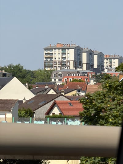 The image shows a residential area with several buildings of varying heights, including apartment buildings and smaller houses with pitched roofs. The buildings are predominantly light-colored, with some featuring red tile roofs. There are trees visible in the background and foreground, some of which are lush and green, suggesting a temperate climate. The sky is clear and blue. In the foreground, a grey, curved structure partially obstructs the view of the buildings, and below it, a section of a wall with graffiti is visible. The graffiti consists of teal-colored shapes on a dark background. The scene appears to be a daytime view from a slightly elevated position, possibly a window or balcony, looking out over a neighborhood. Based on its architectural style and the presence of apartment blocks and houses, this scene is consistent with a residential area in Clamart, France.