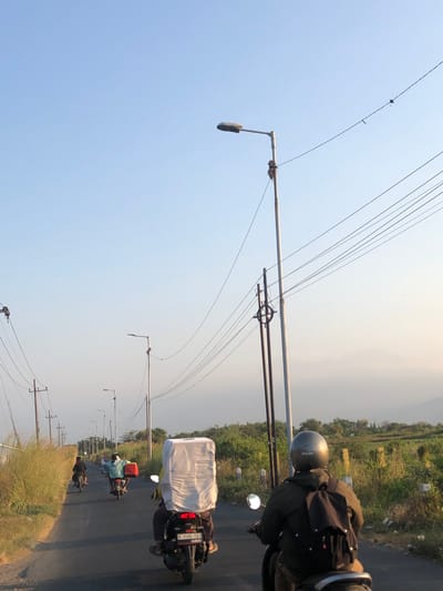 The image shows a paved road with motorcycles driving away from the camera in Glagah Arum, Indonesia. The sky is clear and light blue, with a few wispy clouds in the distance. Several streetlights and utility poles line the side of the road, with power lines stretching across the frame. On the right side of the road, there is a grassy field with some bushes and trees. In the foreground, a person wearing a helmet and a dark jacket is riding a motorcycle, carrying a backpack. Another motorcycle in front of them is carrying a large, white, rectangular object. Further down the road, two more motorcycles are visible, with riders wearing light-colored shirts. The overall impression is one of a rural road in Indonesia during daylight.
