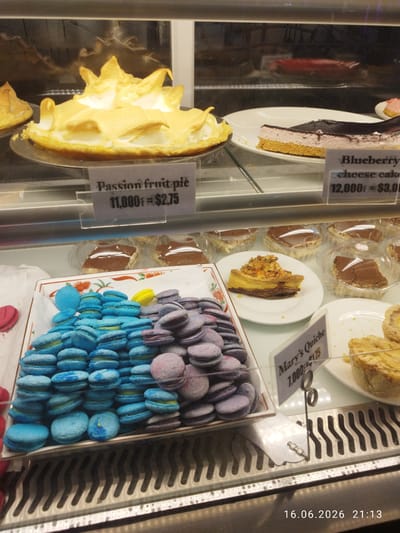 The image shows a display case filled with various pastries and desserts. In the foreground, a tray holds a large quantity of blue and purple macarons, some pink macarons are also visible on the left. Behind the macarons, there are slices of quiche and tiramisu in plastic containers. Further back, a whole passion fruit pie with meringue topping is displayed, along with a slice of blueberry cheesecake and other unidentifiable cakes. A sign next to the passion fruit pie reads "Passion fruit pie 11,000₫ = $2.75". Another sign near the quiche reads "Mary's Quiche 7,000₫ = $1.75". The display case appears to be in a bakery or cafe. The timestamp at the bottom right corner of the image indicates the date and time as "16.06.2026 21:13". The location is identified as Kampot, Cambodia.