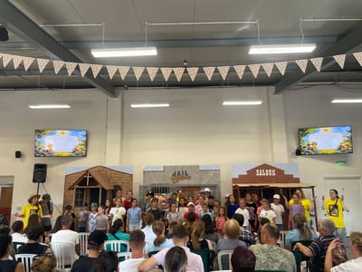 In Zîrnești, Moldova, a group of children are performing on a stage set up in an indoor venue, likely an event hall or community center. The stage features a backdrop with three distinct sections: a wooden house on the left, a building labeled "JAIL" in the center, and a building labeled "SALOON" on the right, suggesting a Wild West theme. A string of colorful triangular pennant flags hangs across the ceiling above the stage.
A group of children, varying in age and gender, are standing in rows on the stage, appearing to sing or act. Two adults are also on stage, one in a yellow shirt to the left, and another in a yellow shirt to the right, both gesturing as if directing or participating in the performance. In the background, a large screen displays an animated image with text in a foreign language, possibly illustrating a story.
The audience, seated in rows of chairs, consists of adults and some children. The environment appears to be a functional indoor space with exposed ceiling structures and fluorescent lighting. The overall scene captures a community event or a school performance.