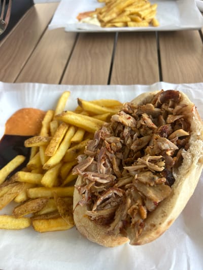 A large, generously portioned meal consisting of a kebab or shawarma sandwich and french fries is depicted in an overhead, slightly angled shot. The sandwich features a split pita bread overflowing with thinly sliced, seasoned cooked meat, likely chicken or lamb, with visible red spice flakes.
A substantial serving of golden-brown french fries is arranged around and partially beneath the sandwich. Two distinct sauces are visible on the white paper lining at the bottom: one light orange/beige and another dark brown/black. The food is presented on white paper, suggesting a casual dining experience, possibly takeaway or fast food.
The setting appears to be a wooden table, with a blurred background showing another plate or container of french fries to the left. The lighting is bright and even, indicating daytime. No people are visible, and there is no discernible text in the image. The scene conveys a hearty, casual meal.