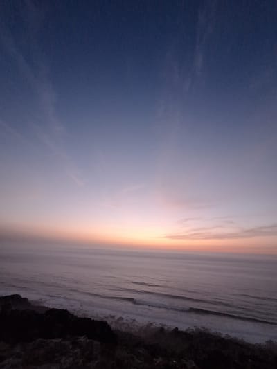 The image captures a serene coastal landscape at dusk, likely near Ahrara, Morocco. The sky transitions from a deep indigo at the top to soft hues of lavender, pink, and orange near the horizon, indicating the sun has recently set. Wispy clouds are scattered across the upper sky, and a faint celestial body, possibly a planet or moon, is visible in the upper left quadrant.

The sea stretches out towards the horizon, its surface marked by gentle, undulating waves that create a subtle texture. The water appears calm, with a muted color palette that reflects the twilight sky. In the foreground, a rocky, dark coastline juts out into the water, with dark, shadowed rocks and a sparse, rough terrain. The overall atmosphere is one of tranquility and natural beauty, characteristic of a coastal sunset. No people or man-made structures are visible in the frame.