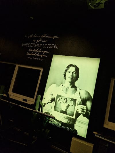 The scene depicts an interior space, likely a fitness-related establishment, located in Oberseelbach, Germany. The environment is dimly lit, featuring dark walls and ceiling, with key elements illuminated by a distinct greenish light. On the upper left wall, a quote is rendered in white script, stating: "Es gibt keine Abkürzungen, es gibt nur WIEDERHOLUNGEN, Wiederholungen, Wiederholungen." attributed to "ARNOLD SCHWARZENEGGER". Below this text, a dark, rectangular opening within a white frame, possibly a window or screen, is partially visible.

Centrally positioned, a large, backlit portrait of Arnold Schwarzenegger is prominently displayed. He is depicted from the chest up, wearing a white ribbed tank top, a watch on his left wrist, and a ring on his left hand. He holds a smaller, framed black-and-white photograph against his chest, which shows him posing in a bodybuilder stance, flexing his biceps.

In the foreground, a piece of dark-colored gym equipment, consistent with a rowing machine or a component of a weight station, extends horizontally across the lower part of the frame. It exhibits metal rails and what appear to be footrests. To the right of this equipment, a light-colored rectangular object, possibly a stand or base, is present. A partial view of another white-framed opening or display is visible on the far right side of the image.