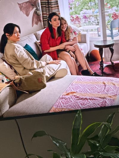 Three people are seated on a sofa in what appears to be a living room. A woman with dark hair pulled back in a ponytail is seated on the left, wearing a tan jacket and beige pants, with a green crossbody bag resting on her lap. Next to her, a woman in a red top is seated, facing away from the camera. Between them, a younger woman with blonde hair, wearing a floral print dress, is seated and leaning on the woman in the red top, smiling and looking towards the camera. Behind them, a large window offers a view of an outdoor area with plants and furniture. The sofa is adorned with cushions and a patterned blanket. In the foreground, a houseplant is partially visible, obscuring the bottom of the image.