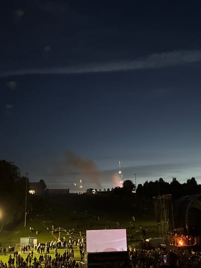 This media file depicts an outdoor event at dusk, likely in Jonava, Lithuania, featuring fireworks and a gathering of people. The sky is a deep twilight blue, transitioning to lighter hues near the horizon, suggesting the early stages of night. Wisps of clouds are visible, and a faint glow emanates from the horizon.

The main activity appears to be a fireworks display, with several bursts of light and smoke visible in the sky. These fireworks are orange and white, illuminating the darkened atmosphere. In the foreground and midground, a large crowd of people is gathered, many of them standing and observing the spectacle. They are situated on a grassy area, likely a field or park, with some individuals positioned behind barriers or in seating areas.

Notable objects in the scene include a large, rectangular screen or stage set up on the right side of the image. It is displaying a light-colored image, possibly a live feed or pre-recorded content related to the event. In the background, there are trees and structures, including what appears to be a tall scaffolding or stage rigging, suggesting a performance or festival atmosphere. A single lamppost is also visible, casting a pool of light.

The overall mood of the image is festive and expectant, characteristic of a public celebration. The gathering of people indicates a shared experience, and the fireworks are a clear indicator of a special occasion. The time of day, dusk, contributes to the dramatic visual effect of the fireworks against the darkening sky. There is no visible text in the media file.
