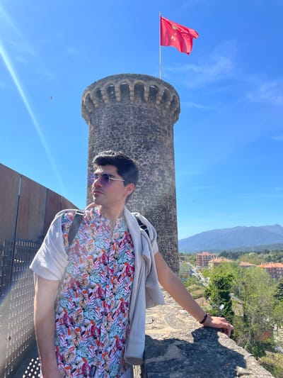 A medium shot features a man in a floral shirt and sunglasses, leaning against a stone structure with his arm extended towards a stone wall. He has a light-colored jacket draped over his shoulder and a dark strap across his body. The stone structure, identified as a tower of the medieval castle in Hostalric, Spain, is to the left and partially behind him. A red flag with a coat of arms flies atop the tower. The background includes a view of rolling hills and buildings. The sky is blue, and a bird is visible in the upper left. There are bright light flares in the lower left corner of the image.