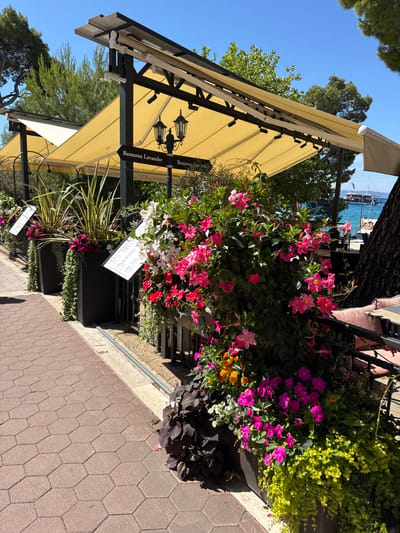 The image shows an outdoor dining area at "Restoran Lavander" in Makarska, Croatia. The restaurant features a yellow awning providing shade over tables. Numerous potted plants filled with colorful flowers, including pink, white, and purple blooms, line the walkway and seating area. A hexagonal-patterned brick pathway leads alongside the planters. In the background, a view of the Adriatic Sea is visible with a boat and some people relaxing on the beach. The overall scene depicts a vibrant and inviting atmosphere on a sunny day.