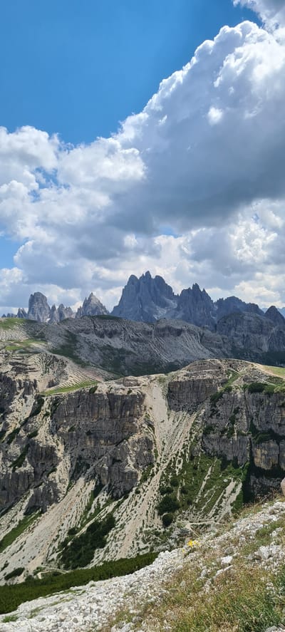 A dramatic mountain landscape unfolds under a partially cloudy sky. The foreground is a rugged, rocky slope sparsely covered with dry grass and a few yellow wildflowers. This slopes down into a steep, terraced mountainside characterized by sheer rock faces, scree slopes, and patches of dark green vegetation. Further in the distance, a range of jagged, grey mountain peaks dominates the horizon, their sharp contours silhouetted against the sky. The sky itself features a mix of bright blue patches and dramatic, gray-white cumulus clouds, suggesting dynamic weather conditions. The scene appears to be a high-altitude, natural environment, possibly part of the Dolomites region near Auronzo di Cadore, Italy, known for its distinctive rock formations. No people or man-made structures are discernible in the immediate landscape, emphasizing the wild and untamed nature of the scene.