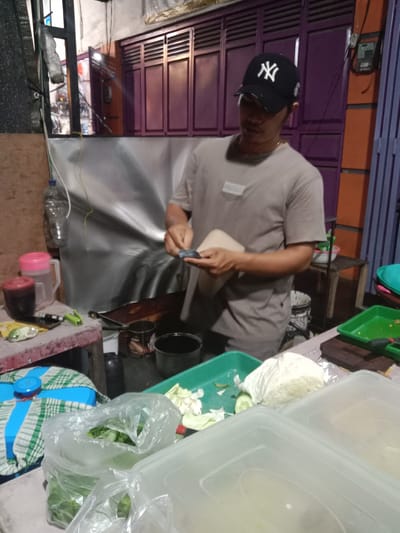 The image depicts a male individual preparing food at a street stall or small establishment in Cangkring, Indonesia. The man, wearing a black baseball cap with a white "NY" logo and a light brown t-shirt with a "PROTEC" patch, is positioned centrally, facing downwards. He is using a small, dark implement in his right hand to work on a light-colored, rectangular food item or wrapper held in his left hand. A thin gold-colored chain is visible around his neck.
To his immediate left, a rustic wooden table holds various items: a red lidded container, a clear plastic pitcher with a pink lid, a green disposable lighter, a shiny metallic object, and a dark, possibly leather, object. A clear plastic bottle hangs from a wire above this table. Behind the table, a silvery-grey tarp or sheet forms a backdrop. Further left, a window shows blurred reflections.
In the mid-ground, a dark frying pan rests on what appears to be a small stove or burner. Several dark pots and a white bucket are visible beneath the work surface.
The foreground on the left features a blue and white checkered cloth covering a round blue container. Beside it are multiple clear plastic bags filled with green leafy vegetables. To the right, white rectangular plastic containers with lids are present. A green plastic tray contains chopped white cabbage and cucumber slices, alongside a dark cutting board and a dark-handled knife. A small, light-colored item wrapped in foil is also on the surface.
The background consists of purple corrugated metal shutters or doors, likely belonging to a building. To the right of these shutters, an orange wall segment is visible, with a grey electrical meter box mounted on it. Various wires are strung across the scene. The overall lighting suggests artificial illumination in an evening or nighttime setting.