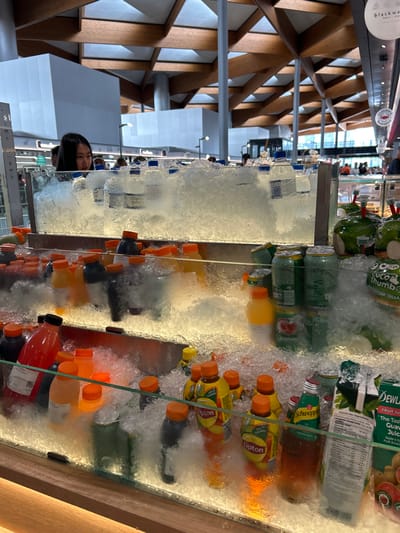 A refrigerated display case is filled with an assortment of chilled beverages on ice. Brands like Lipton, Schweppes, and Dewland are visible. The drinks include juice boxes and bottled drinks in various colors like orange, yellow, and green. Fresh coconuts are also packed among the ice. A person wearing a mask is visible behind the display case. A structure with wooden beams is visible in the background.
