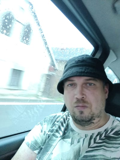 The image shows a man wearing a black bucket hat and a t-shirt with a leaf pattern, looking directly at the camera. He has light-colored eyes and stubble. He is inside a vehicle, likely a car, as indicated by the interior visible in the upper right corner and the window on the left. Raindrops are visible on the window, suggesting it is raining. The background outside the window is blurred, but appears to be a street with buildings. The lighting suggests it is daytime, possibly overcast due to the weather. The man's expression is neutral. There is no visible text in the image. The provided location context is Divišov, Czechia.