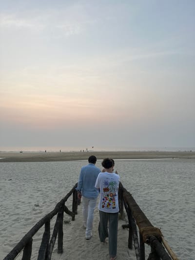 The scene depicts a beach environment in Betalbatim, India, under a sky exhibiting hues of light blue transitioning to pale orange/pink at the horizon. Two individuals are observed walking away from the viewer on a dark, weathered wooden pathway constructed from logs, some wrapped with rope, traversing light-colored sand. The individual on the left, a male, wears a light blue long-sleeved shirt, white trousers, and white shoes. The individual on the right wears a white t-shirt with a multi-colored graphic design featuring palm trees and other elements, and dark green trousers.

The pathway terminates on a wide, sandy beach. Beyond this immediate sandy area, a narrow body of water, possibly a stream or tidal inlet, bisects the beach horizontally. Further in the background, across this water, another expansive sandy area extends towards the ocean. Multiple small, indistinct human figures are scattered across this distant beach, with a red flag visible on a pole, possibly indicating a warning or designated area. The ocean surface appears calm, meeting the sky at a flat horizon. The overall lighting suggests either dusk or dawn.