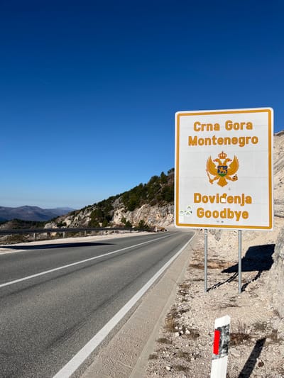 The image depicts a mountainous border area near Klobuk, Bosnia and Herzegovina, under a clear blue sky. In the foreground, a prominent white rectangular road sign with an orange border is visible on the right side of an asphalt road. The sign, supported by two grey metal posts, displays "Crna Gora Montenegro" at the top, followed by the gold Montenegrin coat of arms (a double-headed eagle holding an orb and scepter with a lion on a shield), and "Doviđenja Goodbye" at the bottom. Several small circular stickers are affixed to the lower left portion of the sign. To the immediate right of the sign's base, a white and black reflector post with a vertical red stripe is partially visible in the gravel.
The two-lane asphalt road, marked with solid white lines, curves slightly to the left and ascends into the distance. A concrete curb separates the road from a gravel and rocky shoulder on the right. On the left side of the road, a metal guardrail runs parallel to a steep, rocky mountainside partially covered with green pine trees and sparse vegetation. Distant mountains are visible on the horizon. The scene is brightly illuminated by direct sunlight, casting distinct shadows to the right of the sign and on the left side of the road. A faint airplane contrail is visible in the upper left quadrant of the sky. No people or vehicles are present in the frame.