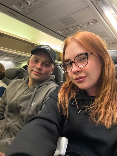 The image depicts two individuals seated inside an airplane. The woman, with long reddish-brown hair and wearing glasses, is positioned at the top of the frame. The man, wearing a black Nike cap and a gray zippered hoodie, is seated below her. The interior of the airplane is visible, with overhead controls and lighting. The location is assumed to be on an airplane departing from Аэропорт Уфа, Russia, based on the prompt.