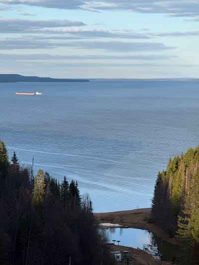 The image shows a body of water with a ship in the distance. The shoreline is visible on the right side of the frame, with trees and vegetation present. A small inlet or channel with boats moored along the side is present in the upper right corner of the image. The sky is cloudy. The location is likely Galevo, Russia.