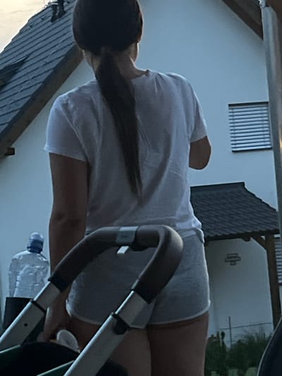A woman, seen from behind, stands in front of a white house with a dark tiled roof. She has dark hair pulled back in a ponytail and is wearing a white t-shirt and grey shorts. In the foreground, a stroller handle and frame are visible. To the left of the woman, a large plastic bottle of water is placed on the ground. The scene appears to be outdoors, possibly in a residential area, with some greenery and a fence visible in the background. The lighting suggests it may be evening.