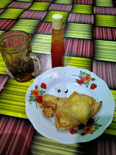 This image displays a dining surface in Lagadar, Indonesia. A patterned tablecloth covers the surface, exhibiting a checkerboard grid of alternating horizontally striped yellowish-green and reddish-brown rectangles. Positioned centrally is a white ceramic plate with a scalloped edge and a floral motif of red and orange roses with green leaves. The plate contains approximately three pieces of light brown, fried food items, one appearing to be a folded pastry or omelet, with some small dark crumbs also present. To the left of the plate, a clear, textured glass mug, featuring a handle and a dimpled pattern, is partially filled with a dark liquid (likely tea), with a small white tag visible submerged within the liquid. Behind the plate and to the right of the glass, a small, transparent plastic bottle with a light yellow flip-top cap stands upright, containing a reddish-orange viscous liquid, presumably a condiment. No individuals are visible.