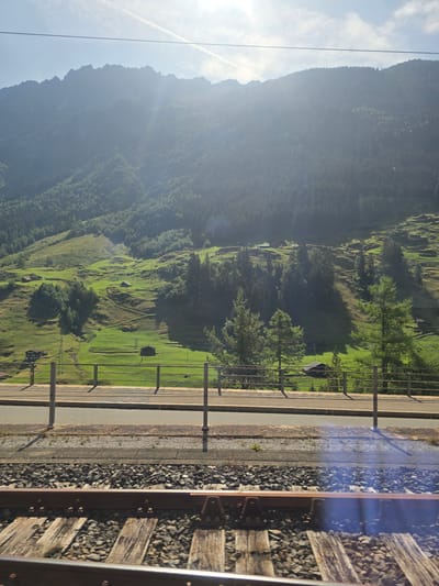 The image depicts a scenic view of a mountainous landscape near the town of Wassen, Switzerland, as seen from a moving train. In the foreground, train tracks with wooden sleepers and ballast are visible, indicating the perspective of a passenger. Beyond the tracks, a metal fence runs horizontally, separating the railway line from a paved area and a grassy embankment. Further in the distance, rolling green hills are dotted with small, dark-colored buildings, likely chalets or farmhouses, interspersed with clusters of trees. The hills are covered in lush vegetation and show the undulations of the terrain. In the background, a large, imposing mountain range rises majestically, its peaks partially obscured by a hazy, sunlit atmosphere. The sky above is a pale blue with some wispy clouds, and the sun is positioned high, casting a bright light and creating lens flare effects on the image. The overall scene conveys a sense of natural beauty and rural tranquility.