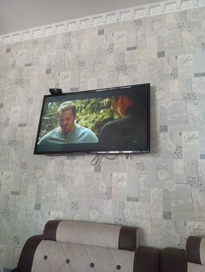 In the city of Chigatay, Uzbekistan, a television is mounted on a wall adorned with patterned wallpaper featuring floral designs and the word "Coffee" repeated. The television screen displays a man with light brown hair, wearing a light blue henley shirt, looking downwards. The man appears to be indoors or in a natural setting with trees visible in the background. Above the television, a small black device with a digital display showing "02:15" is attached to the wall. Below the television, the top portion of a sofa with light beige and dark brown upholstery is visible. The overall scene suggests a living room environment.
