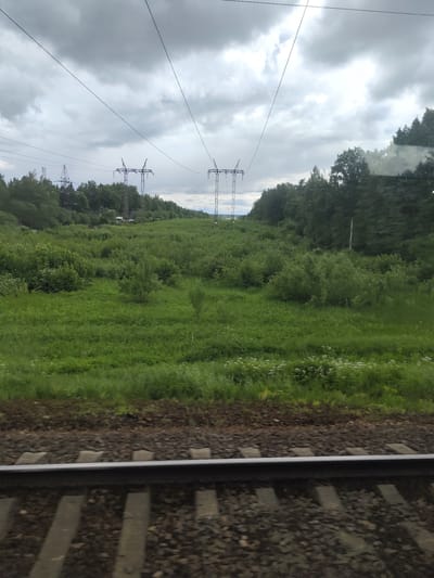 View from a train moving along a railway in Khotkovo, Russia. A green field stretches to a line of trees in the background, with three sets of electricity pylons in the distance. Overhead power lines run above the field. The sky is overcast with grey clouds. The railway tracks are in the foreground.